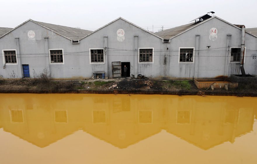 A worker looks at a photographer from a door of a factory manufacturing screws and nuts next to a polluted river in Jiaxing